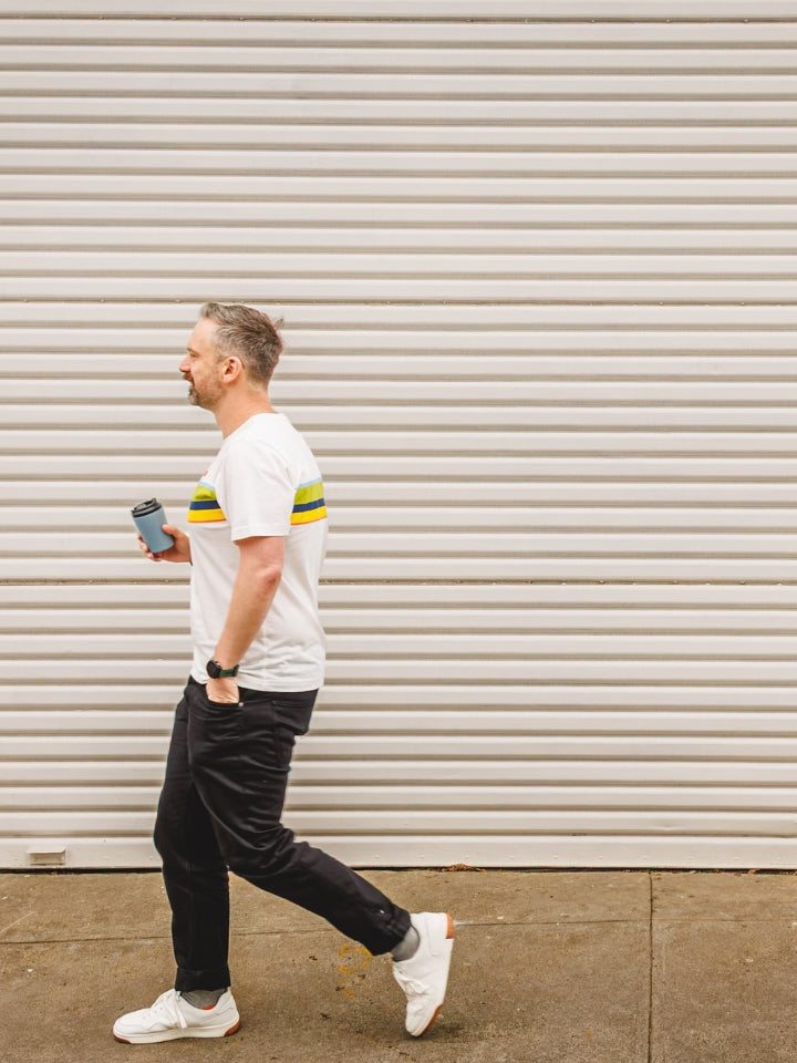 Model walking down a laneway with a LYFER Orange Bellied Parrot Limted Edition t-shirt, holding a coffee with white trainers.