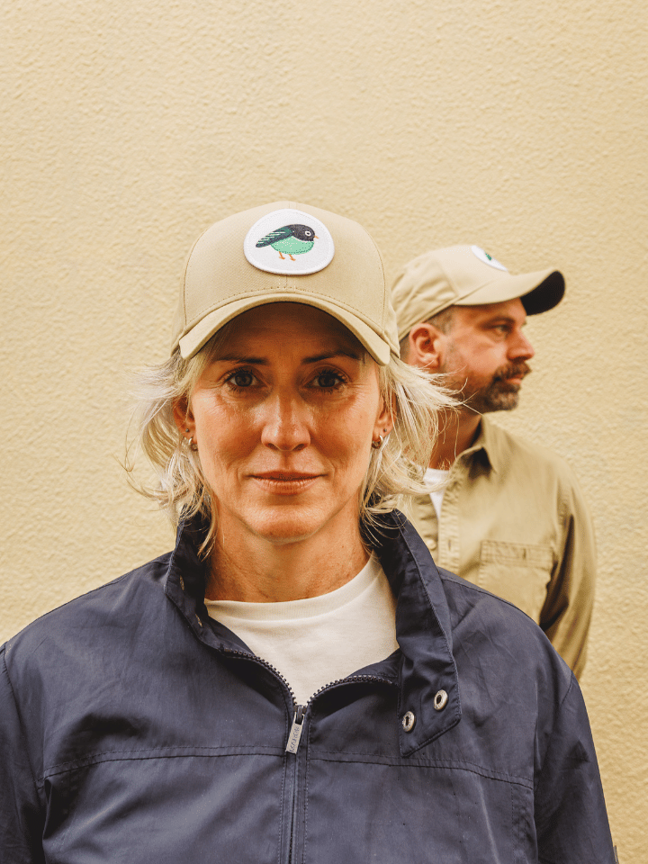 A woman in a navy blue work jacket and beige cap with a bird logo stands in front of a man wearing a matching beige shirt and cap, both against a textured beige wall.