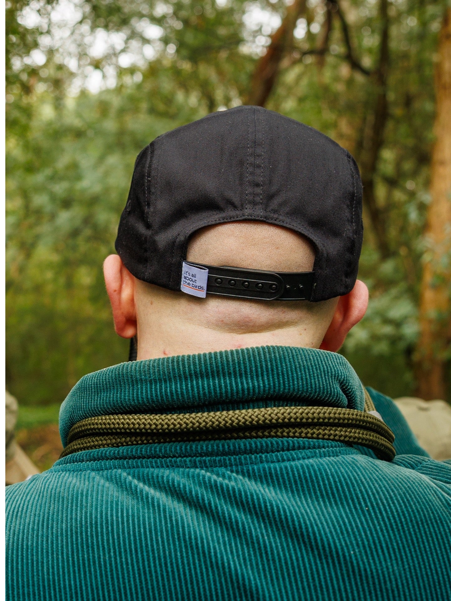 Model wearing Lyfer 5 panel cap with showing the It's all about the birds woven tag on back, while in nature.