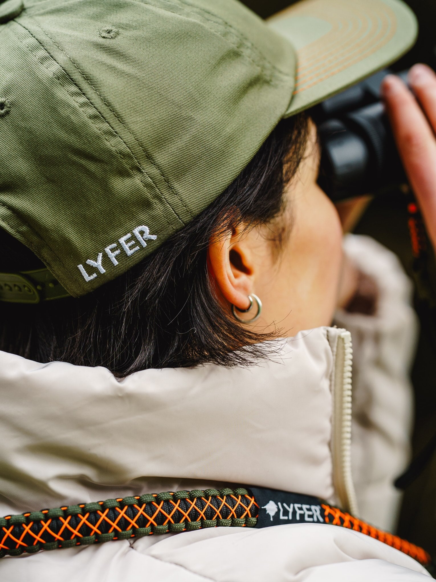 Person wearing a Lyfer green Birdmark 6 panel cap with 'LYFER' branding, looking through binoculars.