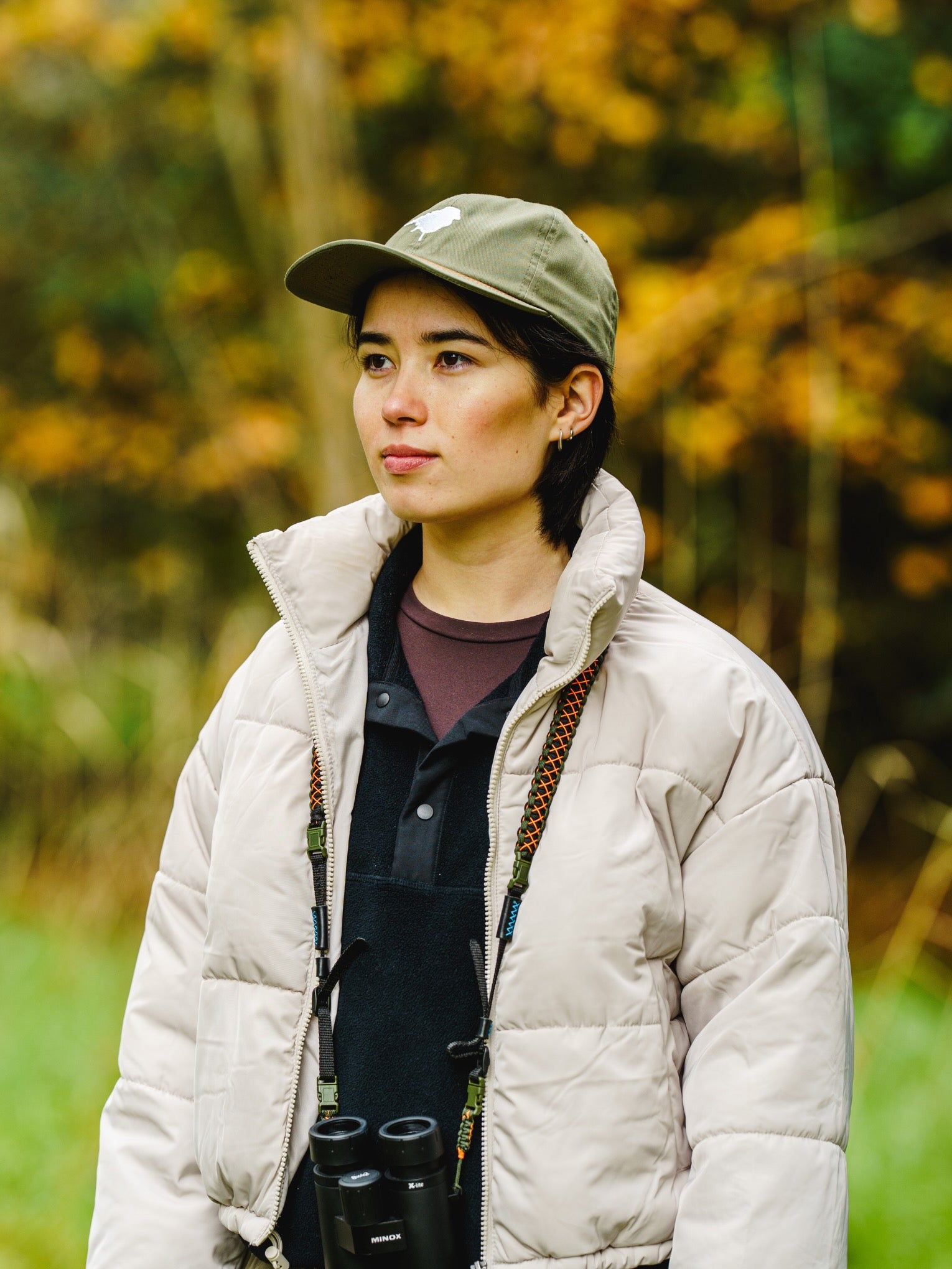 Person wearing a beige puffer jacket and green Lyfer birdmark 6 panel cap with binoculars, standing in a forest.
