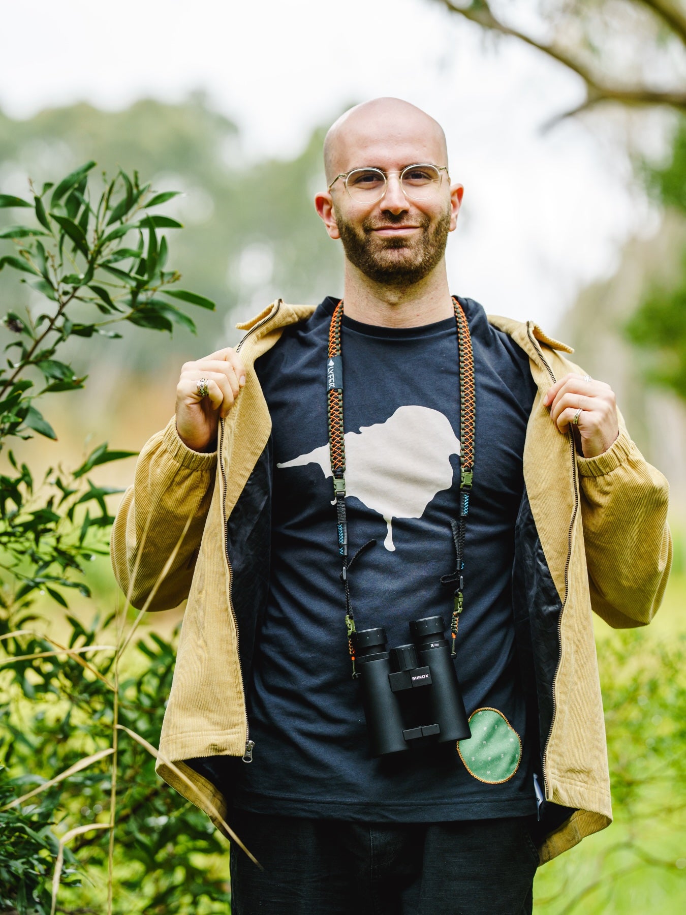 Man wearing a lyfer birdmark t-shirt with a white bird poo graphic, with binoculars, standing in a natural setting