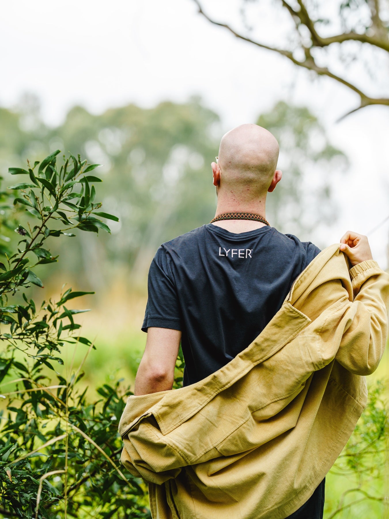 model with outdoor jacket, showing the back of the Lyfer Birdmark T-shirt that says LYFER printed on the back with water-based inks, while out in nature
