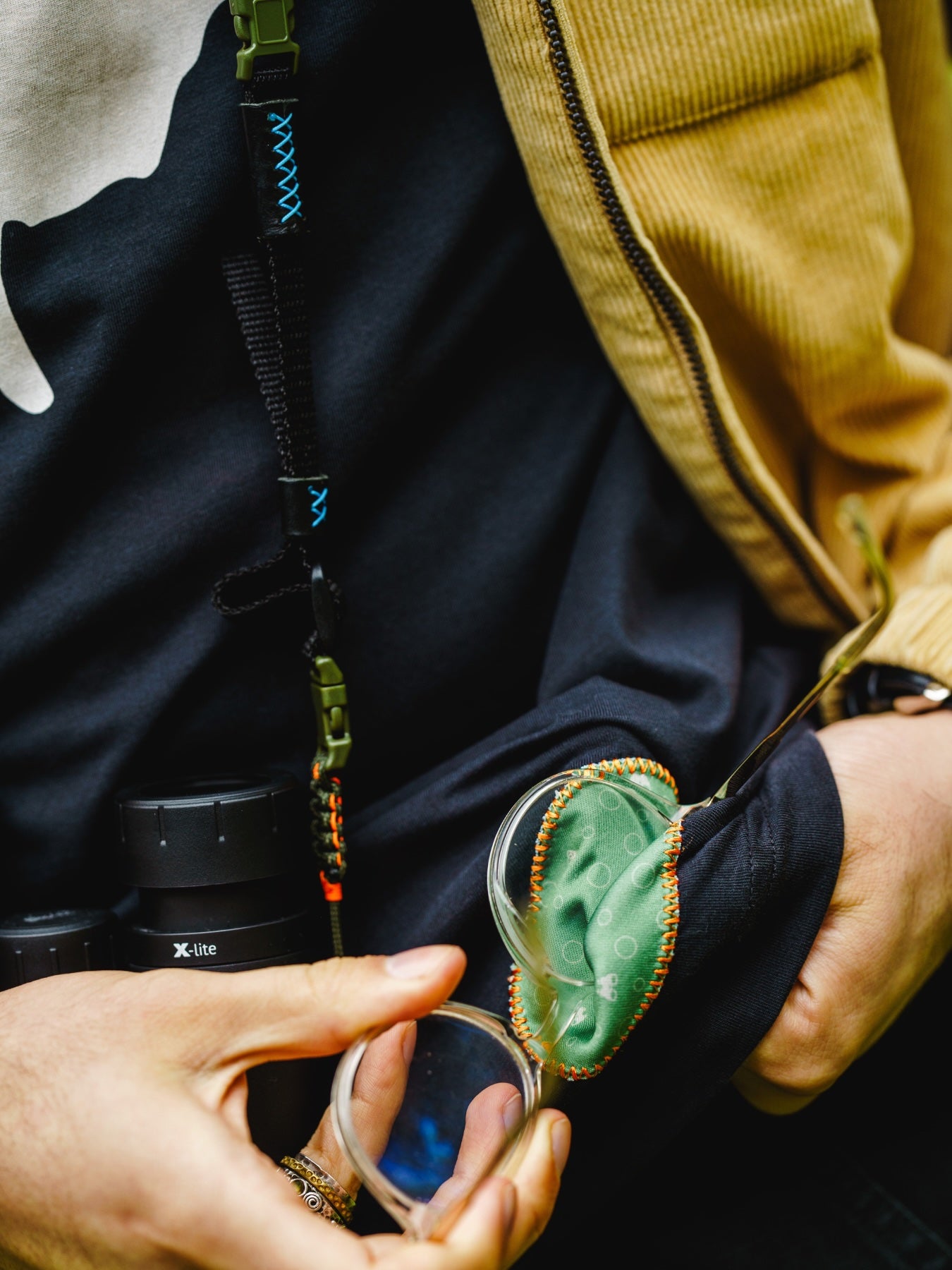 Close up of model using the microcoth binocular cleaner woven into the birdmark lyfer t-shirt, cleaning glasses, while wearing binoculars.
