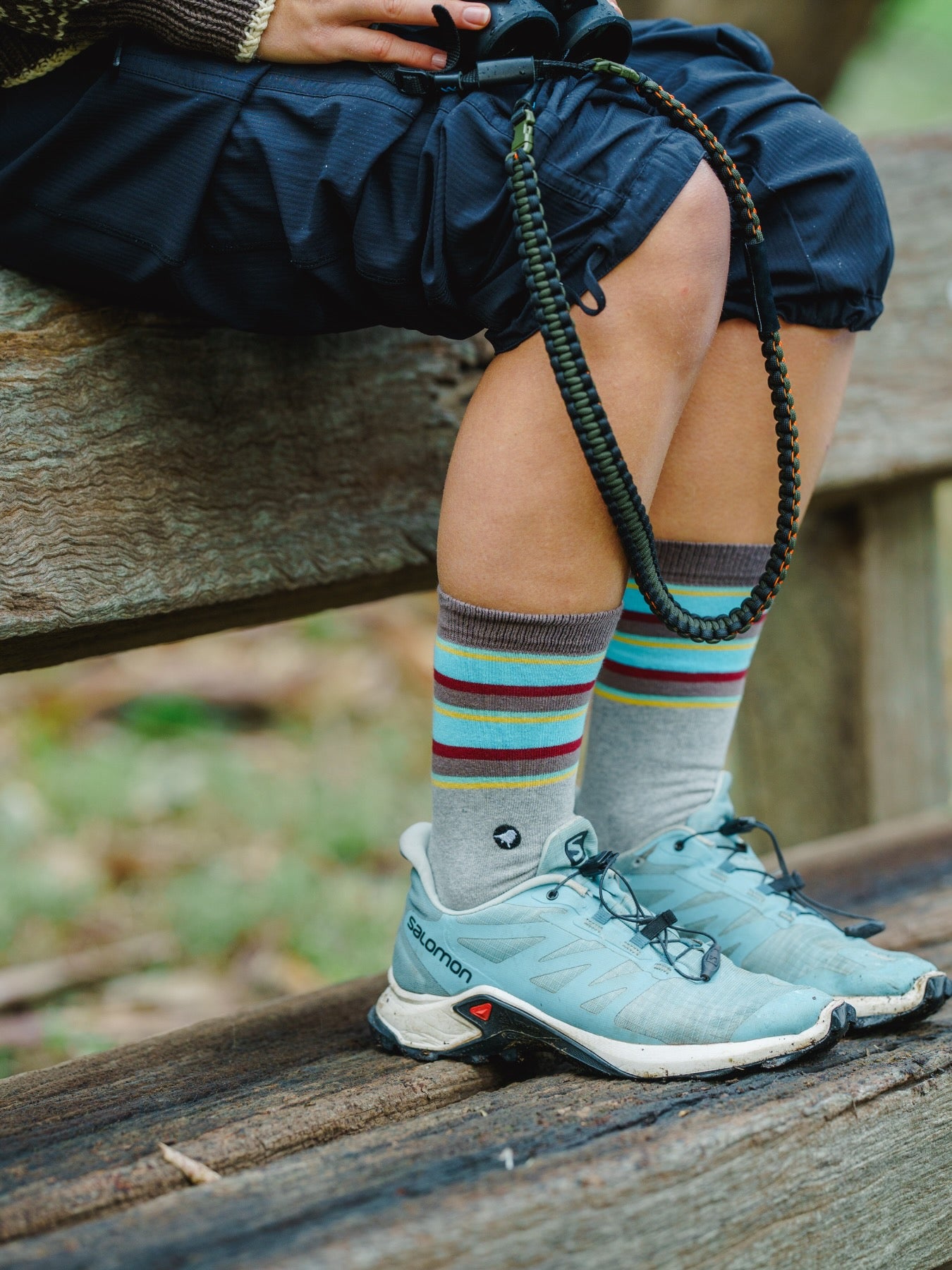 Person sitting on a wooden bench wearing Lyfer Endangered Range socks of the Golden-shouldered Parrot colours in teal hiking shoes.