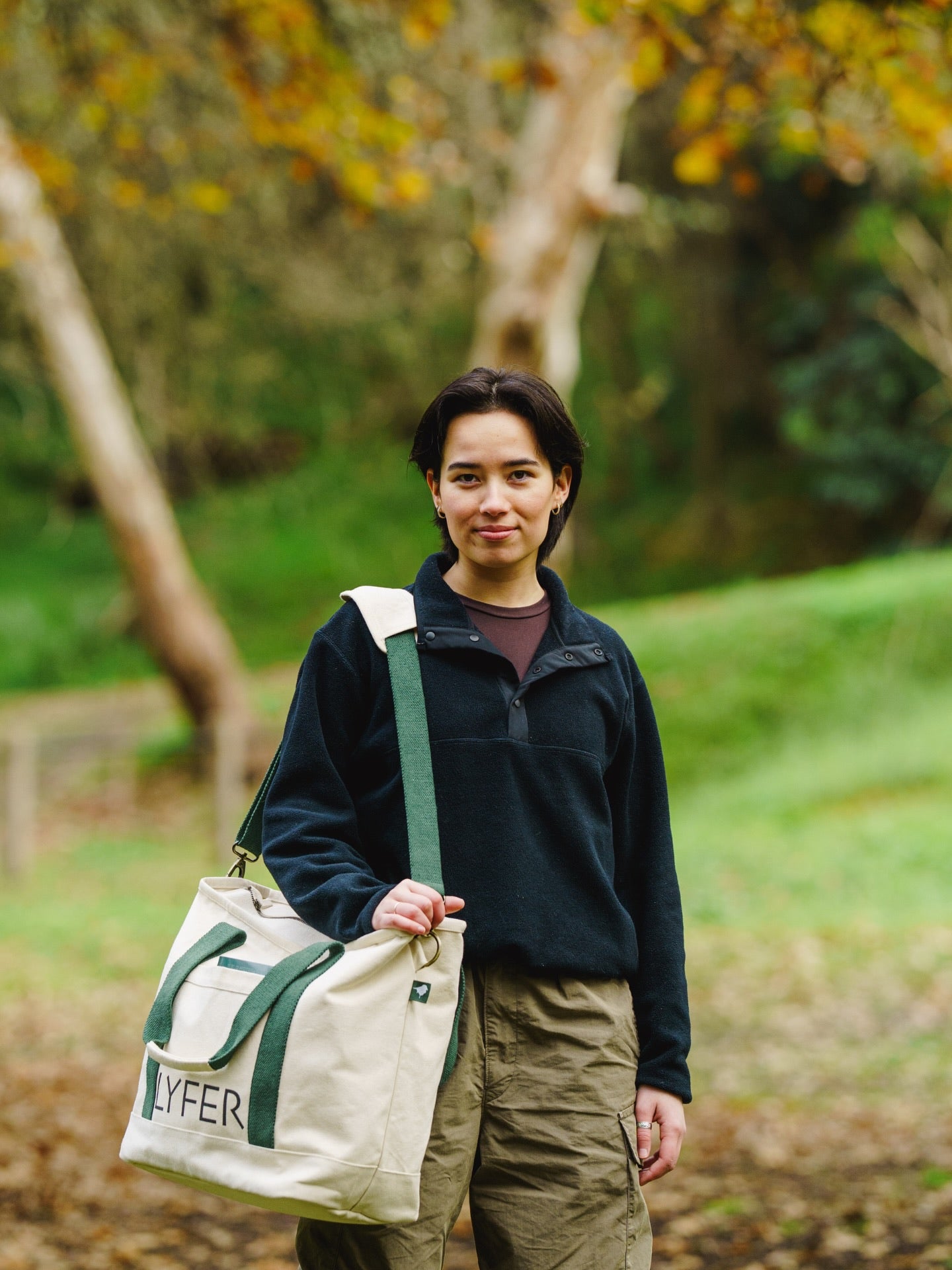 Person holding a Lyfer field tote bag with green straps in a forest setting, with the words LYFER on the bag.