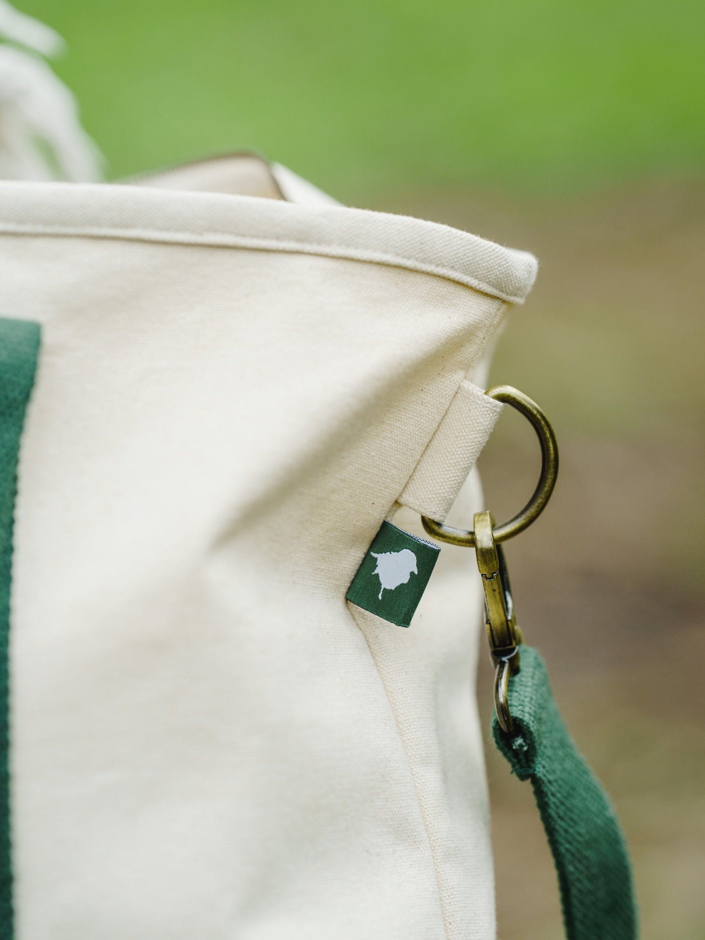 Close-up of a Lyfer field tote bag with green straps and a visible Lyfer birdmark woven brand tag against a blurred natural background.