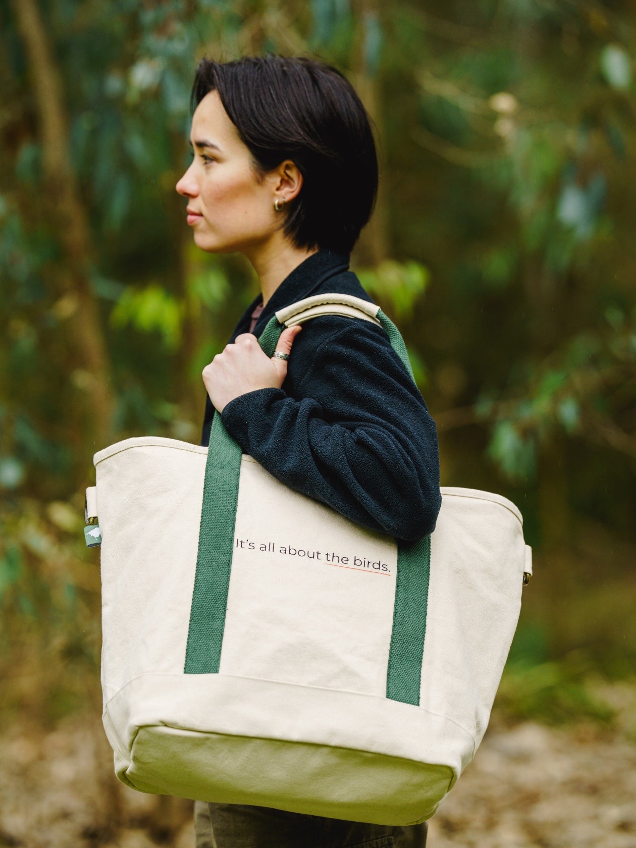 Person holding a Lyfer field tote bag with green straps in a forest setting, with the words It's all about the birds on the bag.