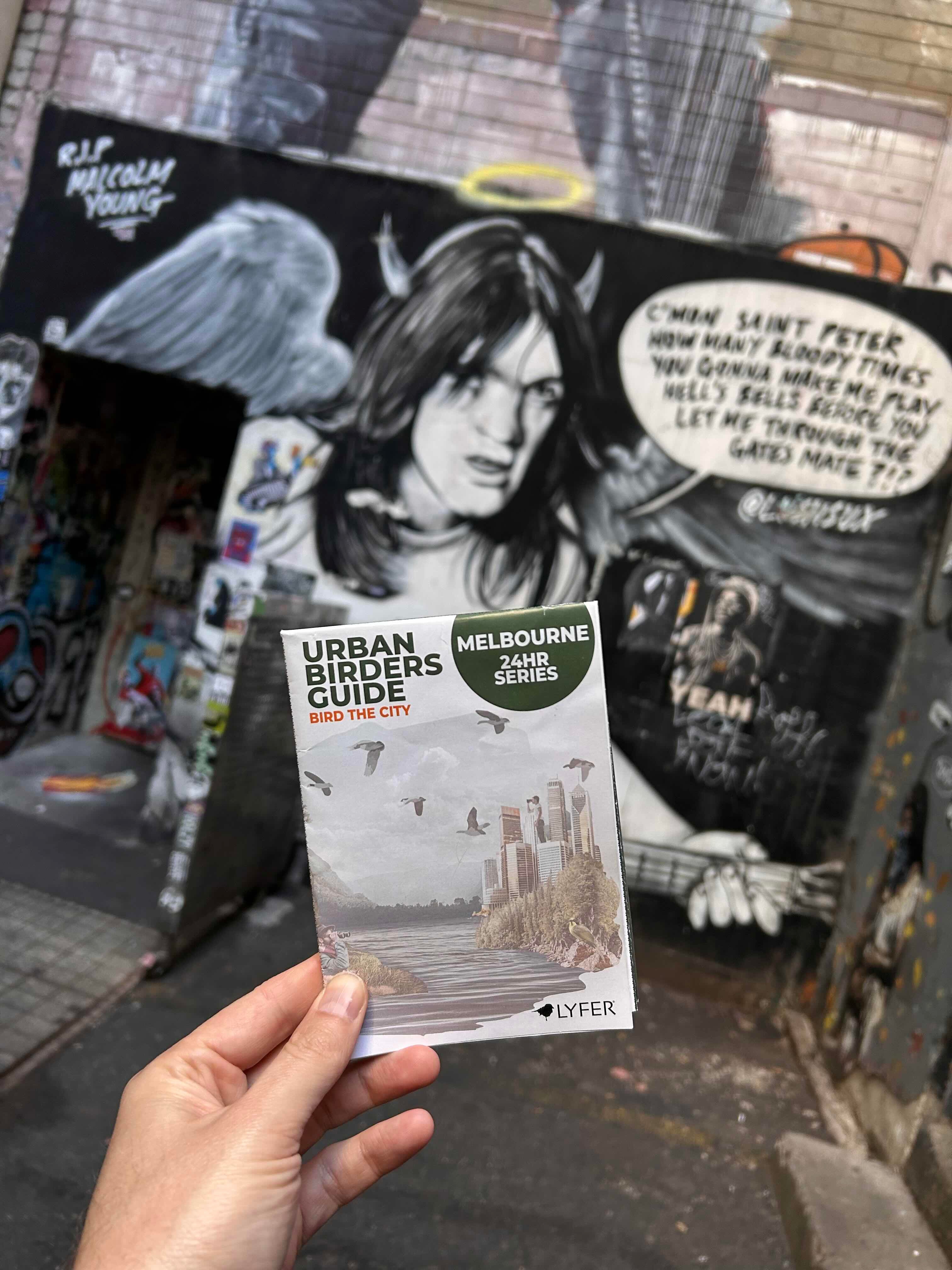 Hand holding a 'Melbourne Urban Guides' leaflet in front of a stylized illustration.