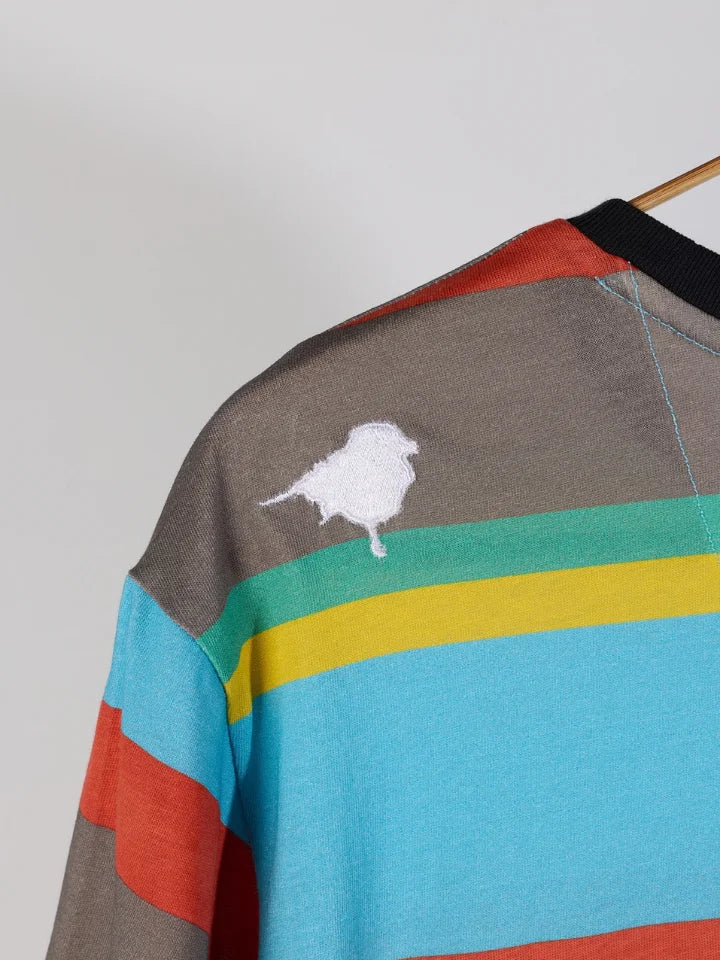 A close-up of a striped knit top on a hanger, featuring horizontal bands of gray, turquoise, yellow, green, and rust red, with a white embroidered bird motif on the shoulder.