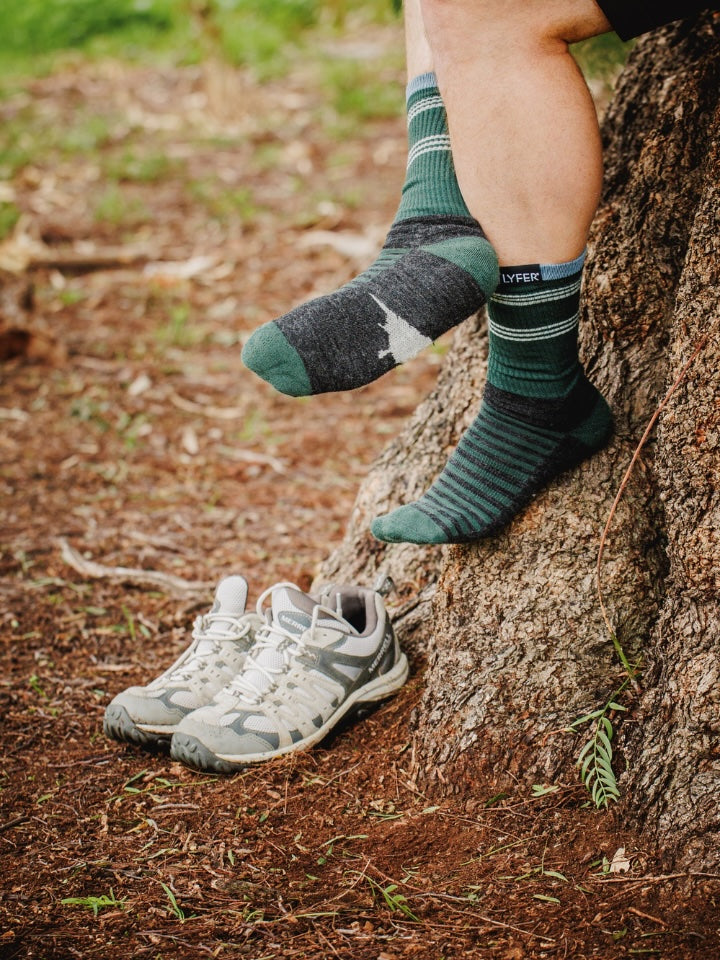 Person wearing green and black striped socks with white sneakers leaning against a tree in a natural setting.