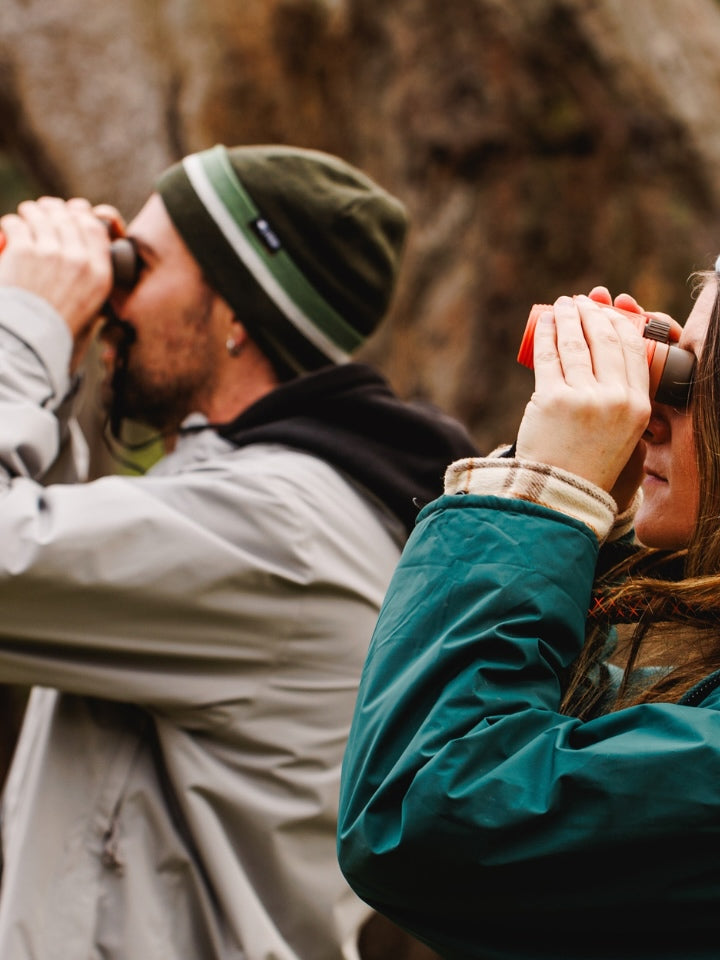 Two models outdoors wearing beanies with  binoculars, looking at birds