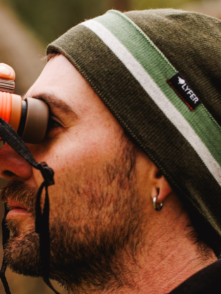Close up of male model wearing LYFER woolie hat made out of merino wool, holding orange NOCS binoculars.