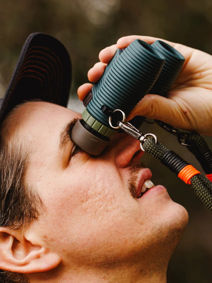A close-up lifestyle shot of a man holding a pair of NOCS Cypress Green ribbed binocular sight to his eye, with a hand adjusting a strap. The binocular sight has a dark gray objective lens, silver metal rings, and an attached black paracord lanyard with an orange accent.