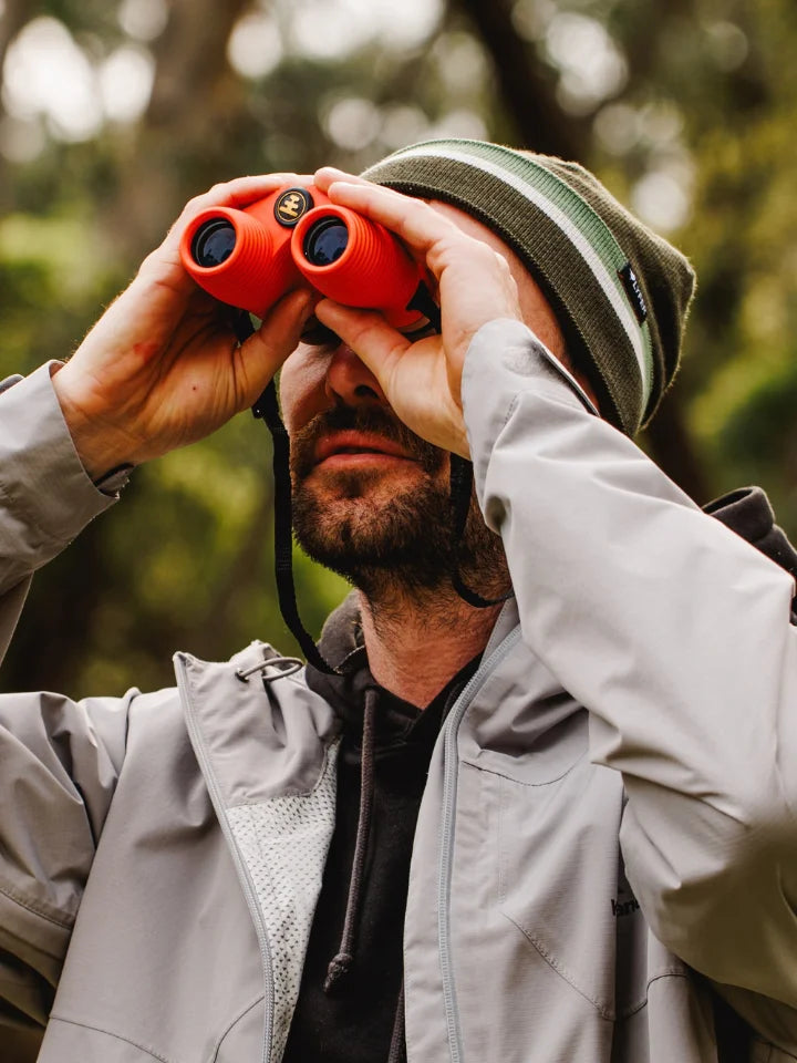 Model holding NOCS binoculars in poppy orange wearing a LYFER beanie looking at birds