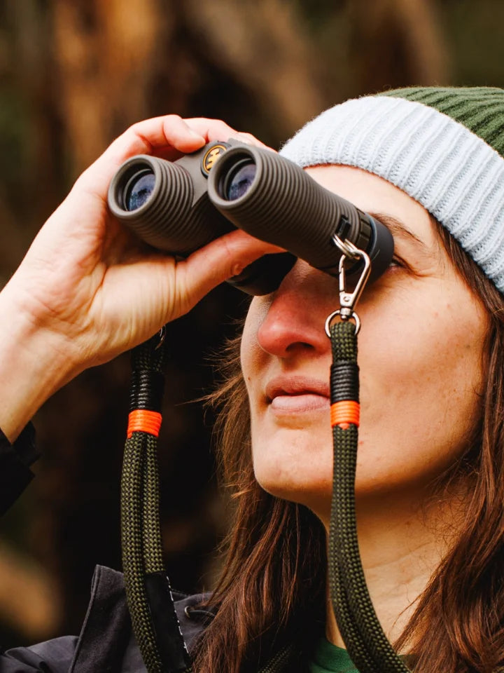 A female model holding NOCS Stone Earth binoculars wearing a LYFER beanie looking at birdse 
