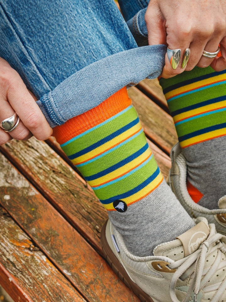 A close-up lifestyle shot of a person sitting on a wooden bench, pulling up the cuff of their blue jeans to reveal a pair of colorful striped socks. The socks feature horizontal stripes of orange, yellow, green, and blue, worn over gray crew socks and paired with beige sneakers.