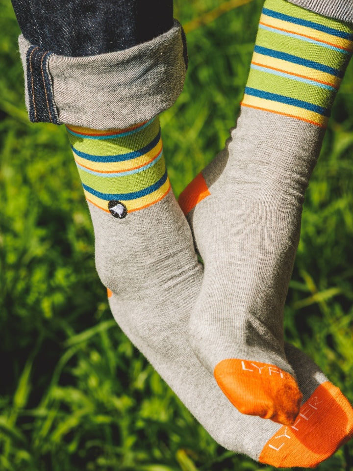 A close-up of a person's feet wearing gray crew socks with bright green, yellow, and blue stripes, orange toes, and dark denim cuffed at the ankles, set against a blurred green grass background.