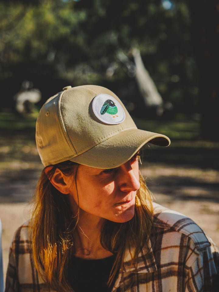 A woman wearing a tan LYFER Patch cap with a circular bird patch on the front, photographed outdoors in warm sunlight with a blurred natural background.