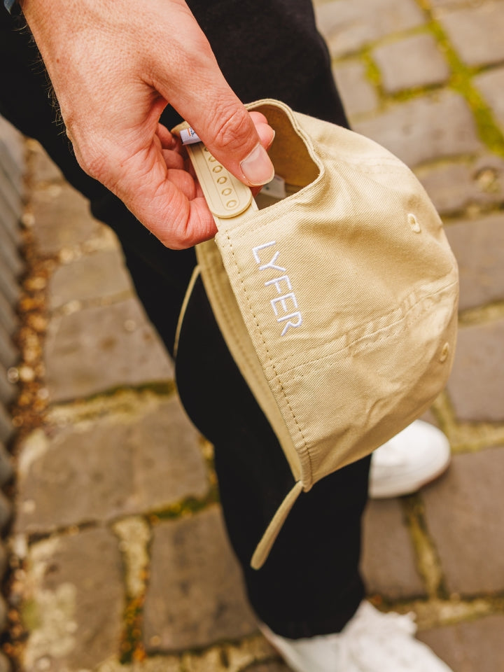 A close-up of a hand holding a beige baseball cap with the brand name 'LYFER' printed vertically in white on the side, set against a blurred background of a person's legs and white sneakers on a stone pavement.