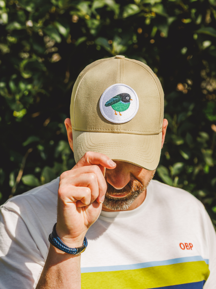 A man outdoors wearing a beige baseball cap with a circular embroidered bird patch on the front, paired with a white t-shirt against a background of green foliage.