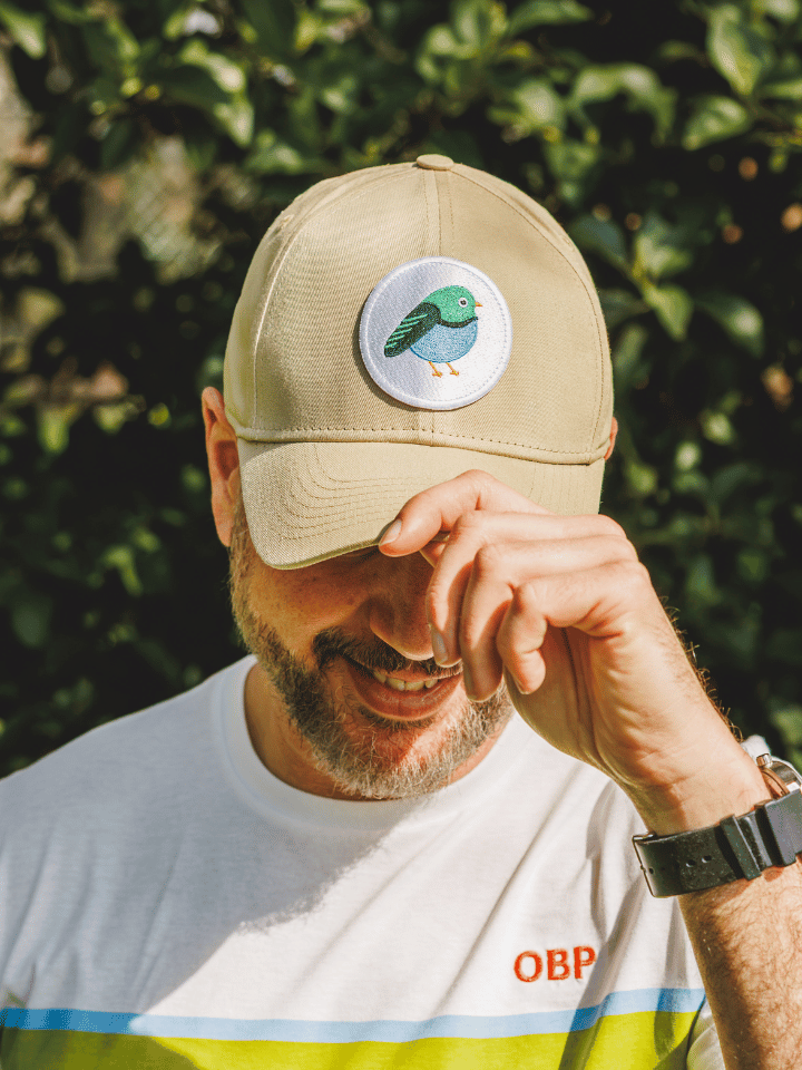 A man outdoors wearing a beige baseball cap with a circular embroidered bird patch on the front, paired with a white t-shirt against a background of green foliage.