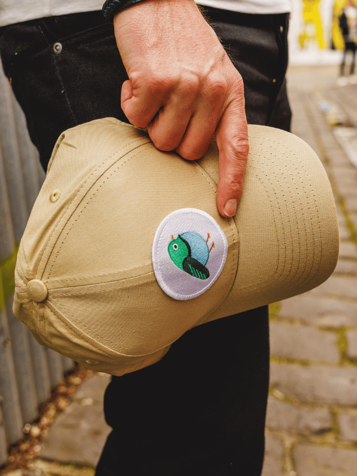 A person holding a beige baseball cap with a circular embroidered bird patch on the side, set against an outdoor background.