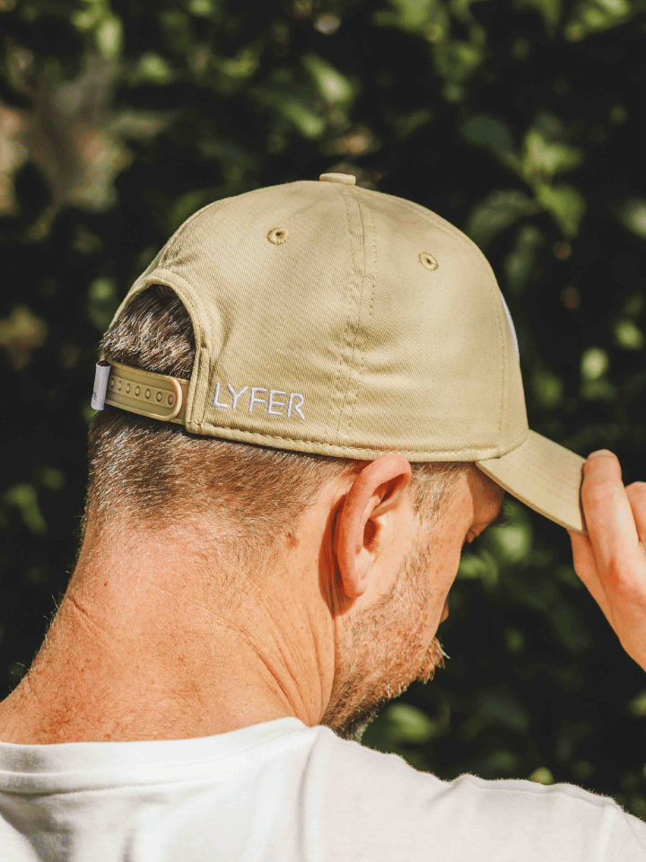 A man wearing a beige baseball cap with 'LYFER' embroidered on the side, photographed outdoors against a blurred green foliage background.