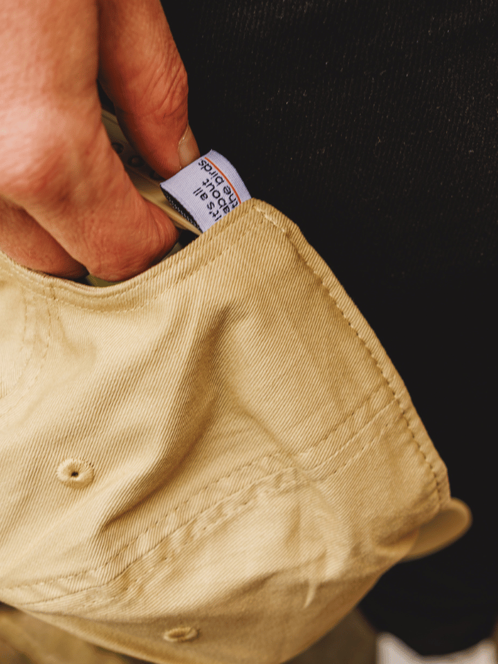Close-up of a hand pulling a white brand tag from the pocket of a beige canvas cap, with a black shirt in the background.