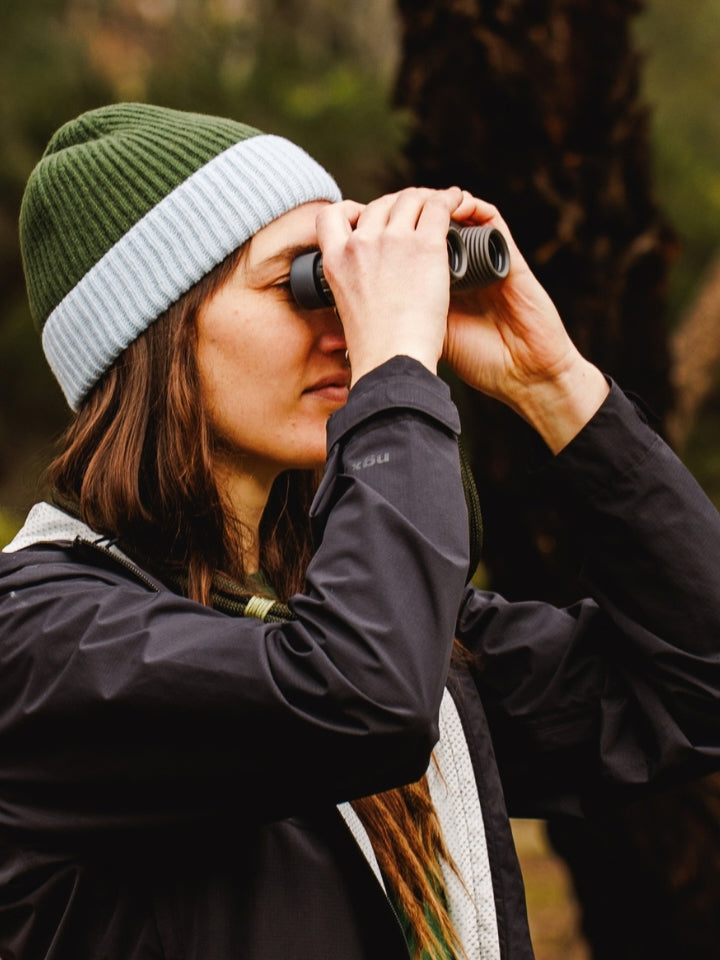 Female model outdoors holding binoculars looking for birds, wearing a ribbed knit wool beanie in green and blue