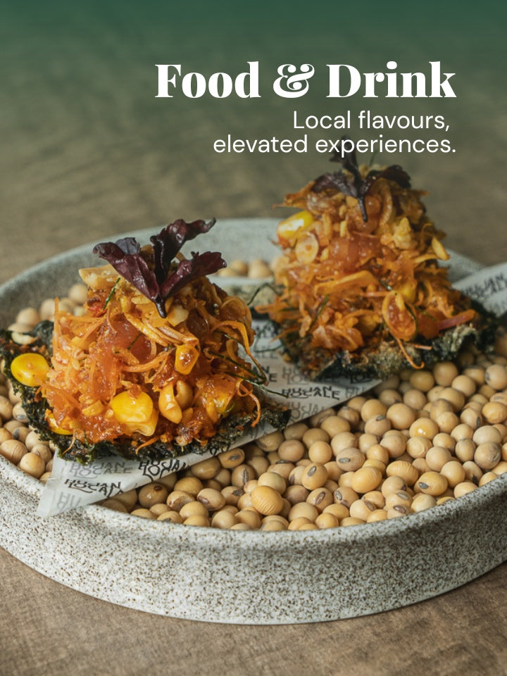 Two gourmet stuffed flatbreads topped with shredded orange sauce, corn kernels, and greens, served on a speckled stone plate filled with soybeans. The background is a rustic wooden surface with a green gradient overlay and the text 'Food & Drink Local flavours, elevated experiences.