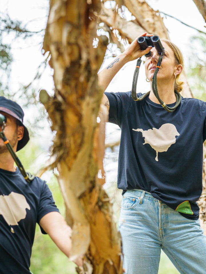 Two models in a tree wearing the Lyfer Birdmark t-shirt looking through binoculars looking at birds. 