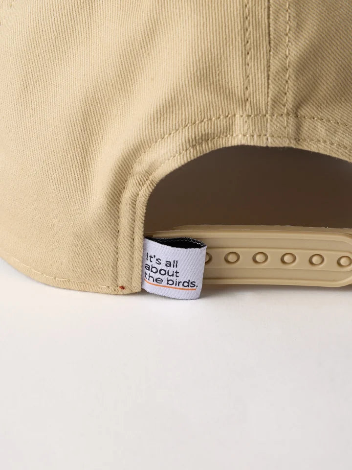 Close-up of the back of a beige baseball cap showing the adjustable strap and a small white fabric label that reads 'It's all about the birds.'.