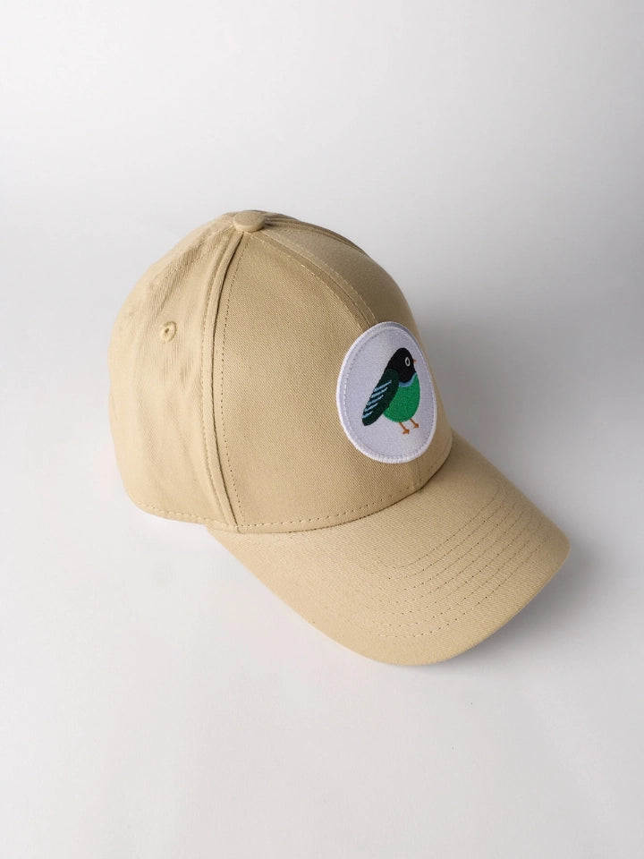 A beige baseball cap with a circular white patch on the front featuring a stylized bird in black, green, and white, photographed on a light gray background.