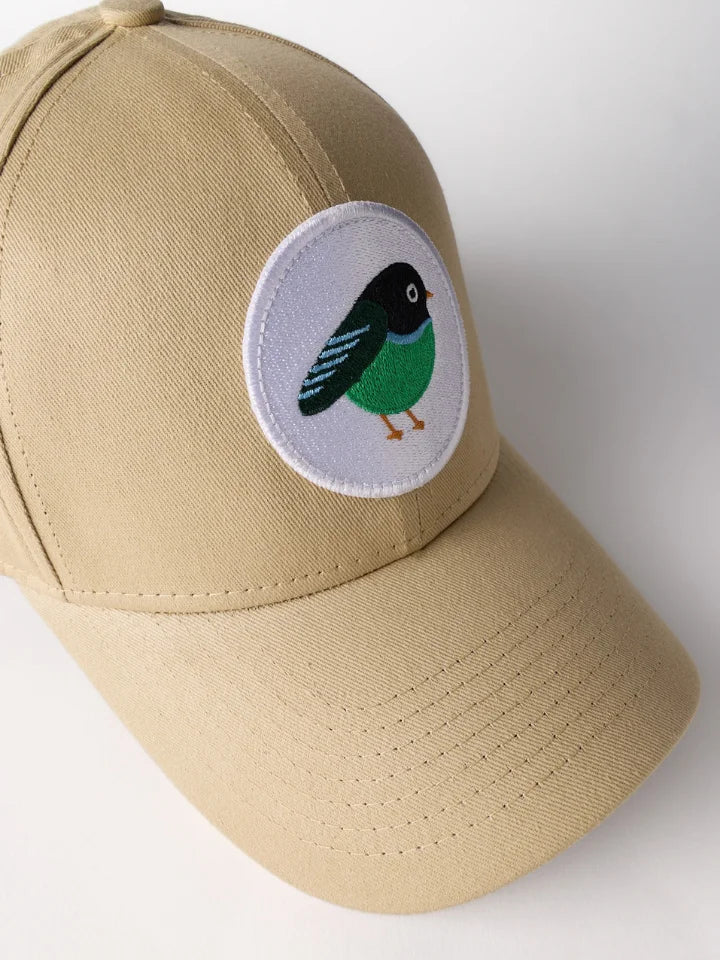 A khaki baseball cap featuring a circular embroidered bird patch on the front. The patch depicts a small bird with a black head, green body, and blue wings on a white background.