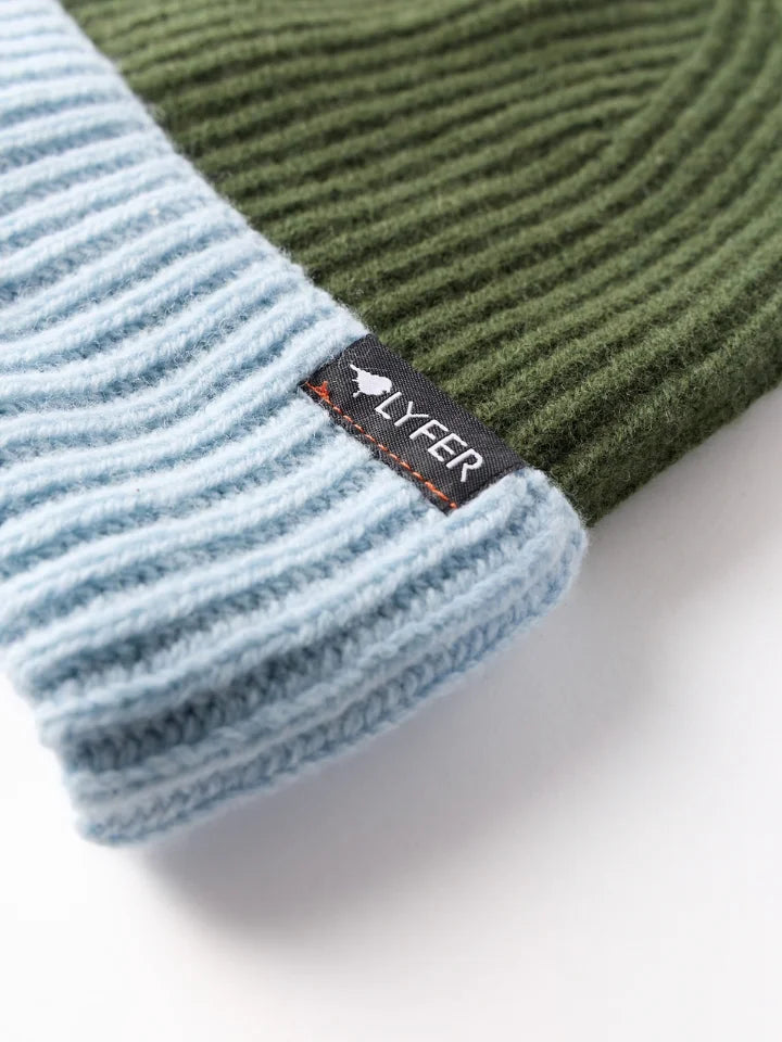Close photo of a ribbed knit beanie hat with an olive green top section and a light blue folded cuff, featuring a small black brand LYFER tag on the side.