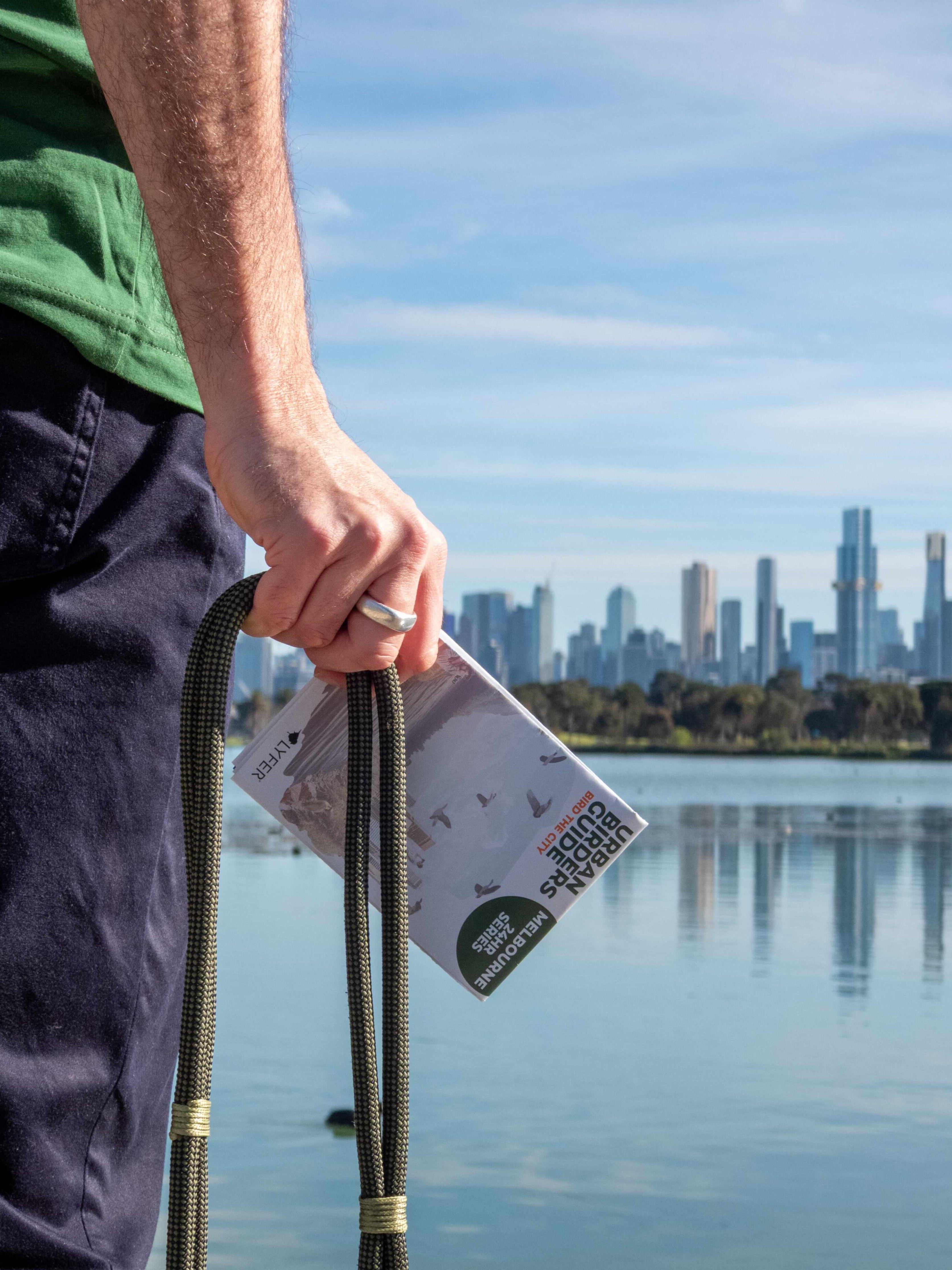 Person holding a binoculars and the Urban Birders Guide by Lyfer ope with a scenic city view of melbourne in the background